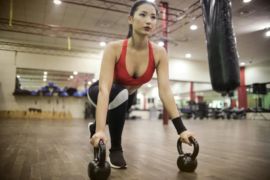 Dari bawah fokus atlet wanita muda Asia dalam pakaian olahraga jongkok dengan kettlebell sambil berlatih sendirian melawan interior kabur gym modern yang ringan