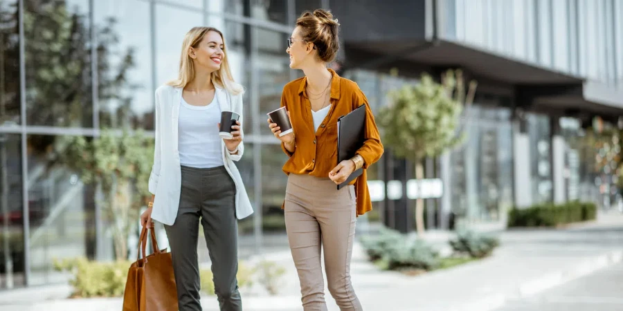 Ganzkörperporträt von zwei jungen Geschäftsfrauen, die mit Kaffeetassen in der Nähe des modernen Bürogebäudes im Freien spazieren gehen