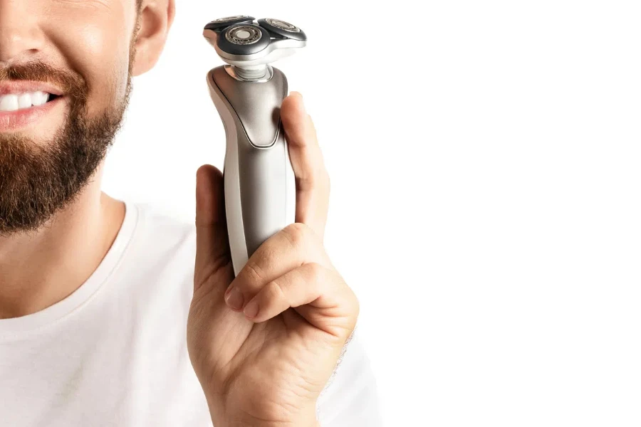 Guapo hombre barbudo sosteniendo una afeitadora eléctrica en su mano sobre fondo blanco.