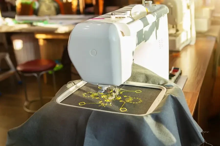 Taller de costura en casa, puesto de trabajo frente a la ventana, con una moderna máquina de bordar que crea un patrón amarillo sobre una tela de lino gris. Al lado del hilo y la tela