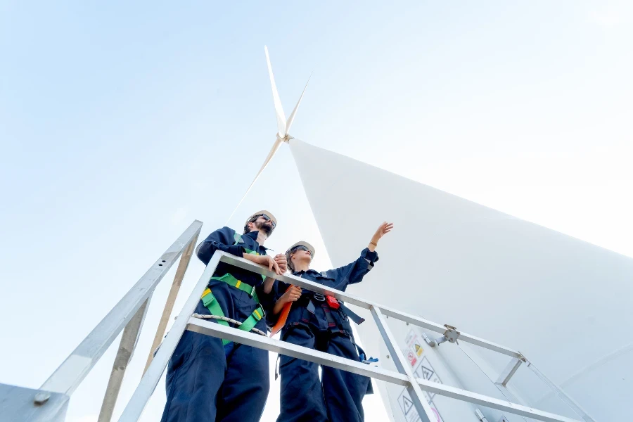 Lower view of professional technician man and woman stay on base of windmill or wind turbine and woman also point to the right with blue sky