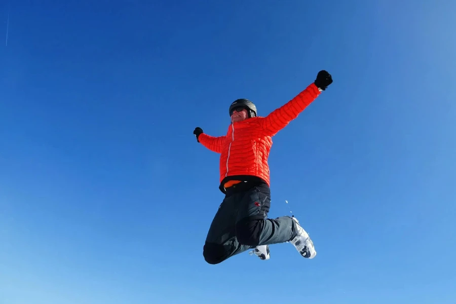 オレンジ色のジップ ジャケットと黒いパンツを着た男が晴れた青い空の下でジャンプ