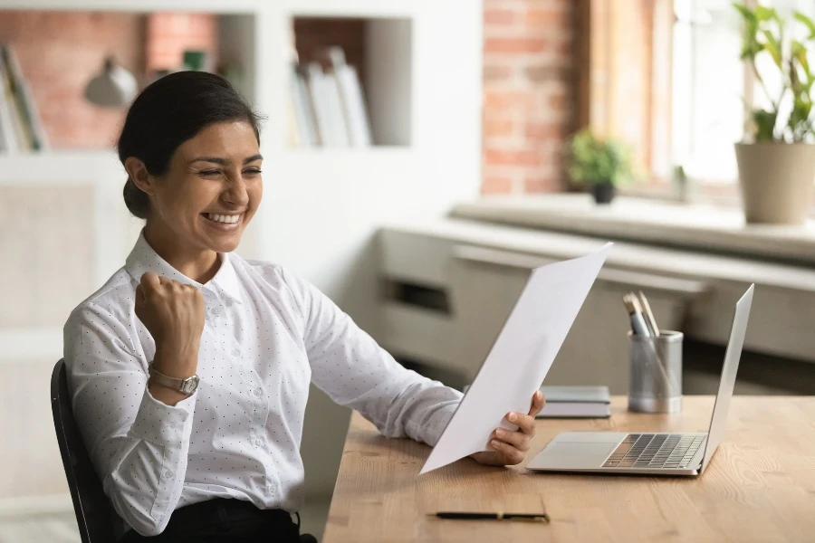 Femme d'affaires indienne souriante et ravie lisant de bonnes nouvelles dans une lettre, assise au bureau, une jeune femme excitée a reçu une bourse, une promotion d'emploi ou une offre, montrant un geste de oui, célébrant le succès