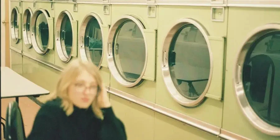 Perspective view of modern washing machines in laundry room with blurred unrecognizable female in eyeglasses sitting at table in black clothes by Rafael Gonzales