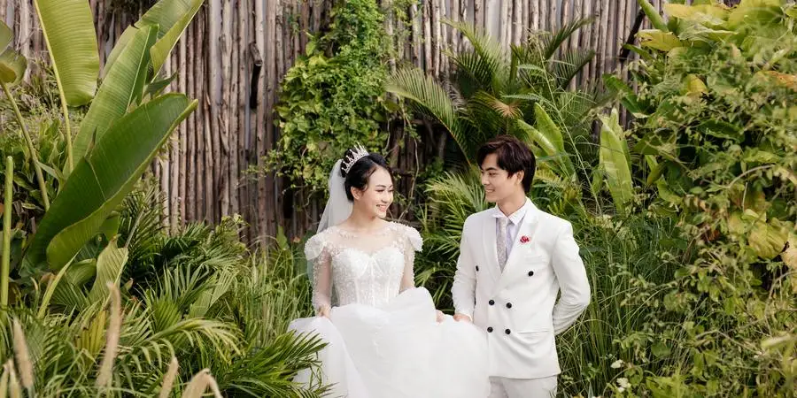 Positive Asian newlywed couple standing in greenery by Jin Wedding