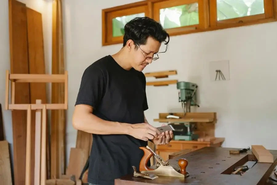 Side view of concentrated male craftsman standing with planer knives at wooden workbench with jointer during work in professional studio by Ono Kosuki