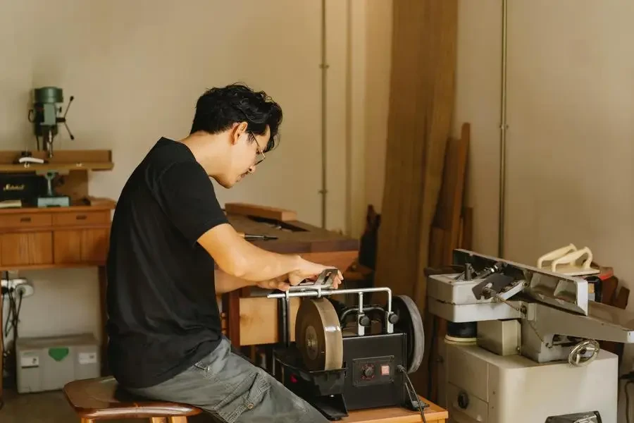 Side view of young concentrated Asian male joiner working on grinder with sharp wheels while sitting on stool in workshop by Ono Kosuki