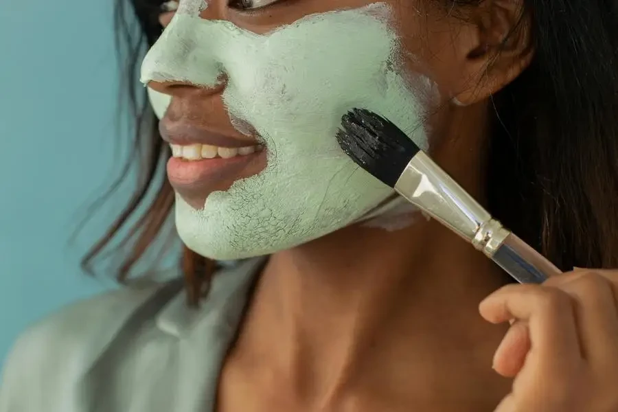 Smiling ethnic woman applying mask on face with brush by Monstera Production