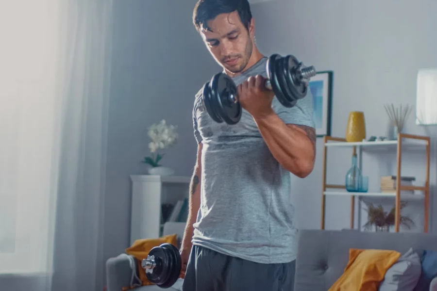 Крепкий спортивный мужчина в футболке и шортах делает упражнения по поднятию икр с гантелями дома в своей просторной и светлой квартире с минималистичным интерьером.