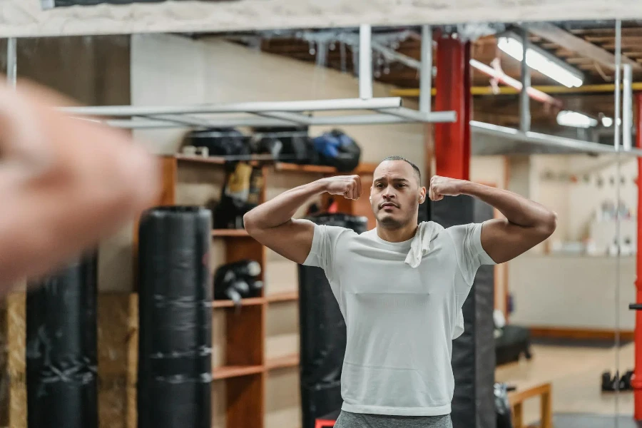 Deportista étnico fuerte mostrando bíceps y reflejándose en el espejo del gimnasio