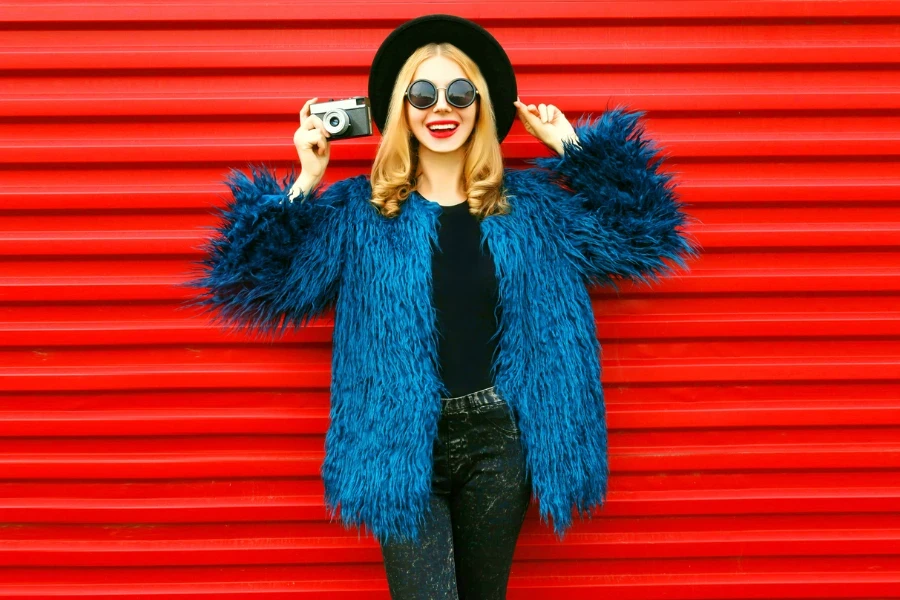 Elegante modella sorridente con macchina fotografica retrò che indossa un cappotto di pelliccia sintetica blu