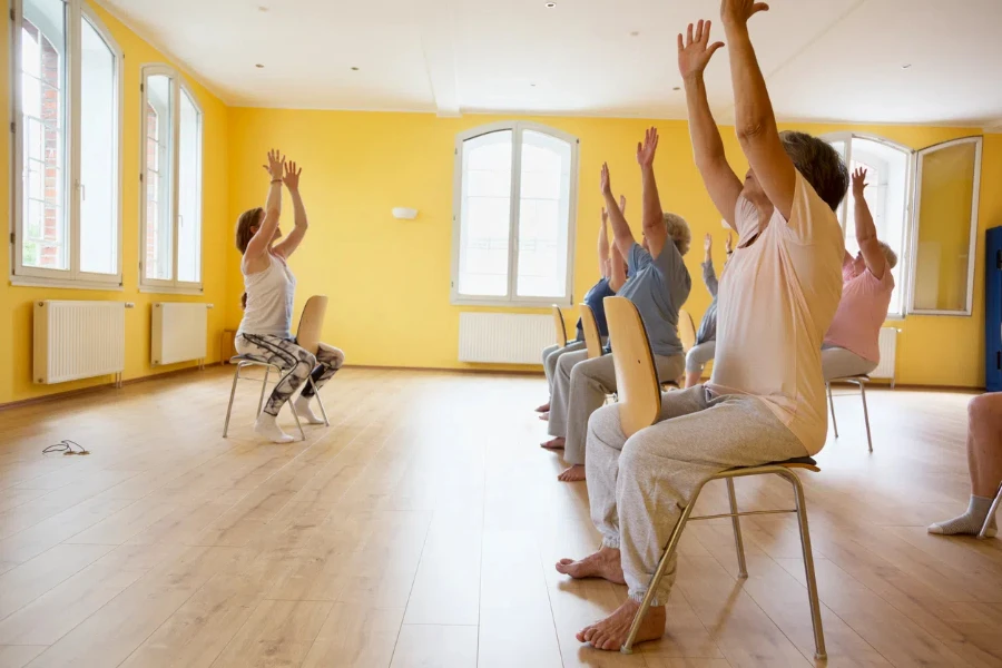 Professora e aulas ativas de yoga para mulheres idosas em cadeiras, braços levantados