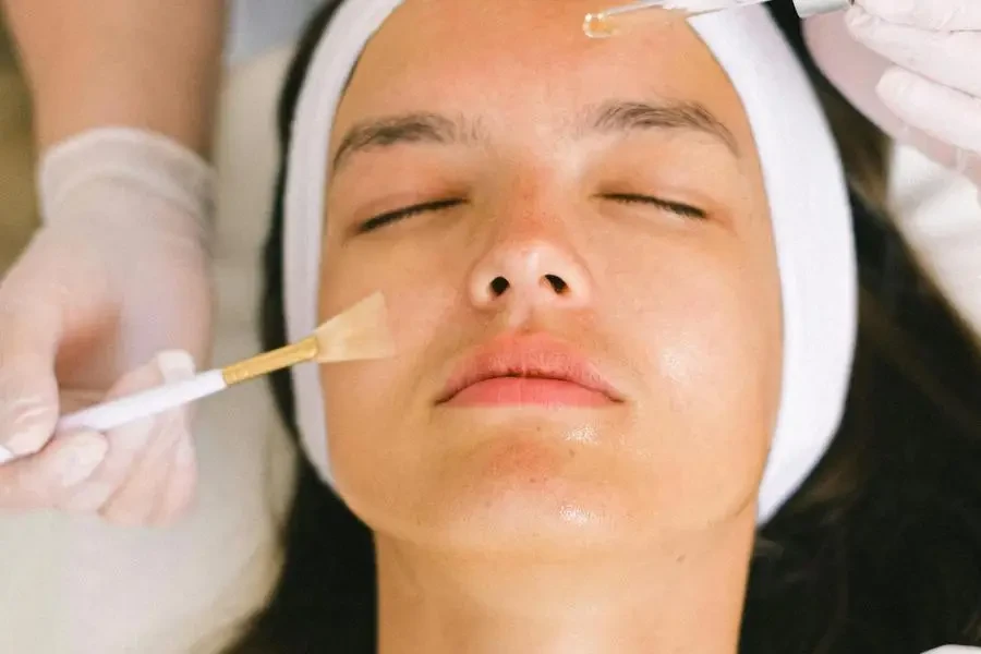 Top view skilled female cosmetologist in uniform and gloves applying essential oil on young woman face skin while working in contemporary spa center by Anna Shvets