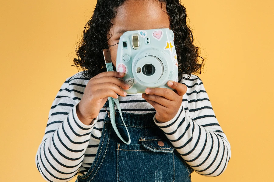 Unrecognizable little stylish ethnic girl in denim jumpsuit taking photo on contemporary instant photo camera while standing against brown background