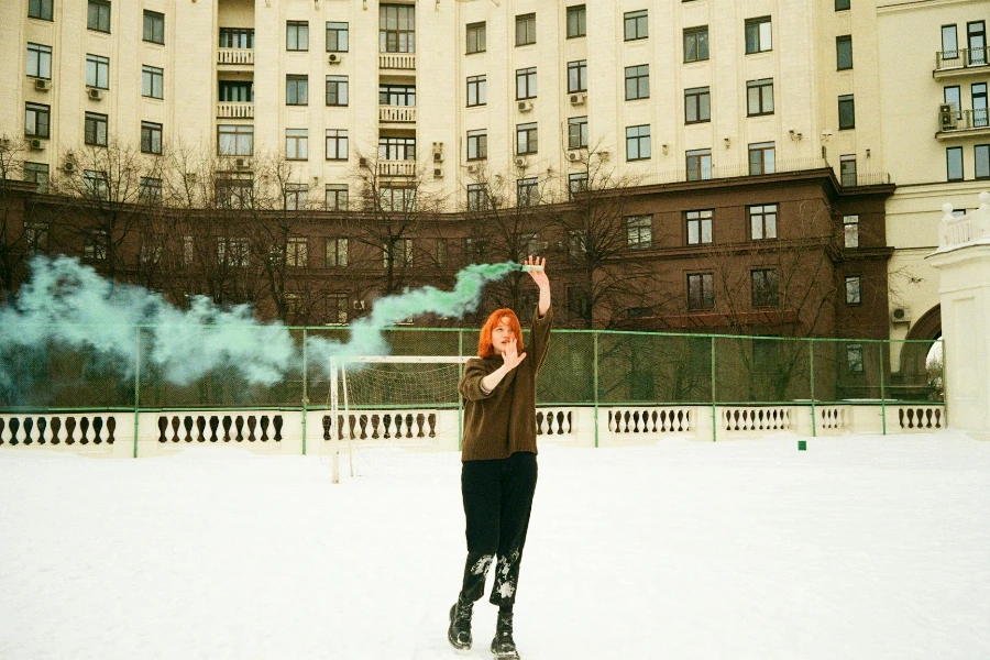 Femme tenant une fusée de fumée verte au-dessus de sa tête sur un terrain de sport scolaire couvert de neige