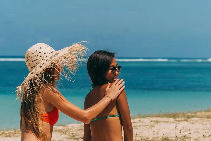 Donna con cappello da sole e bikini rosso che applica la lozione sulla schiena di una donna in bikini verde di Mikhail Nilov