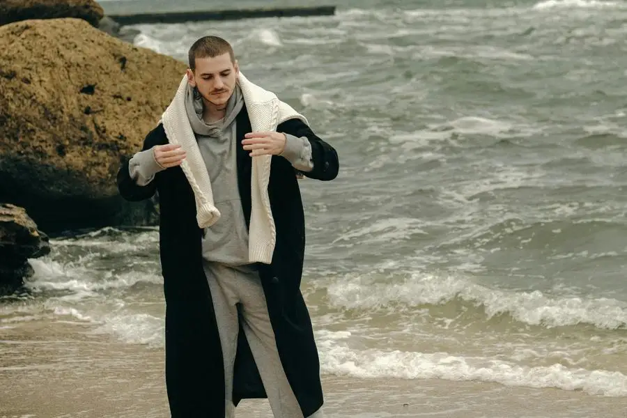 Joven de pelo corto con ropa deportiva y abrigo largo y cálido posando en la orilla del mar por Valdemoro