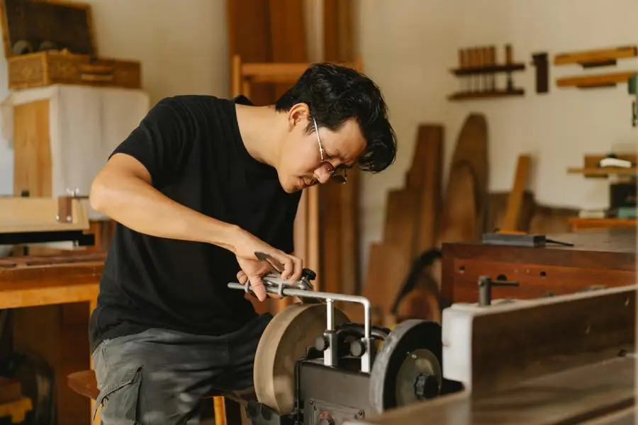 Young attentive ethnic male joiner in eyewear sharpening manual instrument on grinder with wheels in light workroom by Ono Kosuki