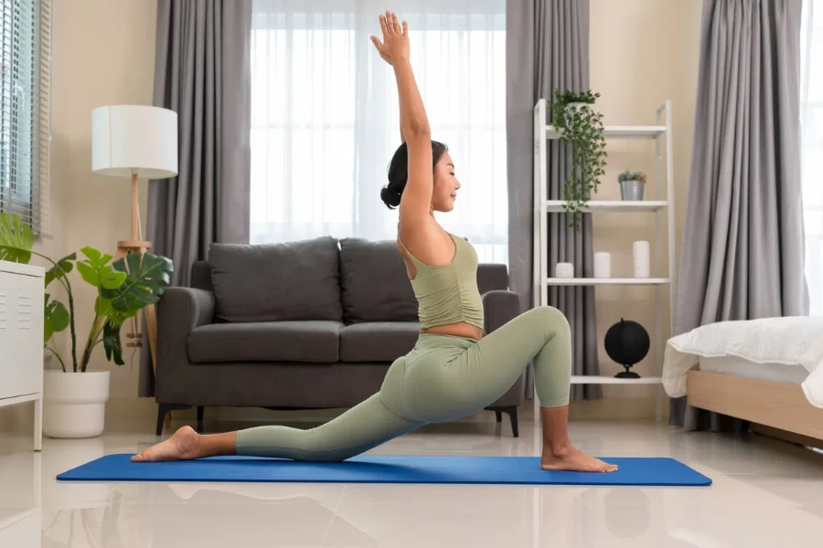 Wanita muda mengenakan pakaian olahraga hijau dan berlatih yoga pagi secara rutin di rumah, berdiri dalam pose Low Crescent Lunge atau Anjaneyasana untuk meregangkan paha dan selangkangan di atas tikar biru di kamar tidur