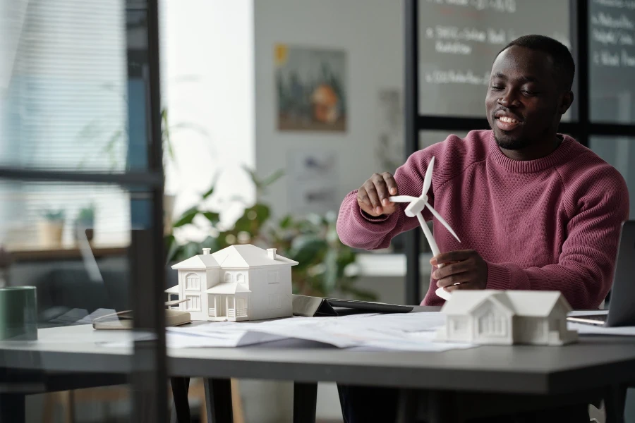 Un jeune architecte souriant fait tourner la turbine d'un modèle d'éolienne tout en étant assis sur son lieu de travail avec un plan déroulé et une maquette de maison