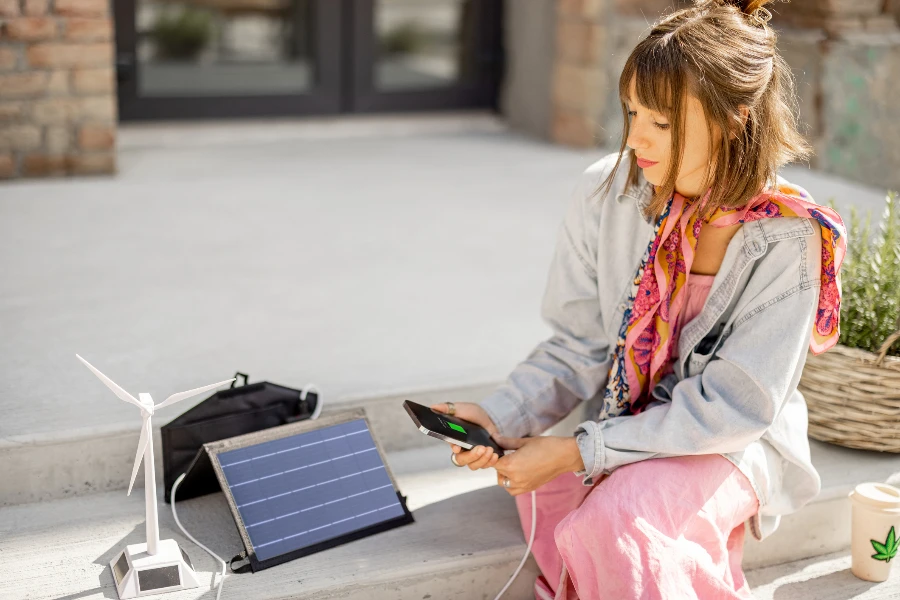 Una giovane donna elegante usa lo smartphone e lo carica dal pannello solare portatile mentre è seduto rilassato sulla strada all'aperto