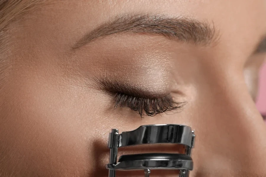 Young woman using eyelash curler on pink background