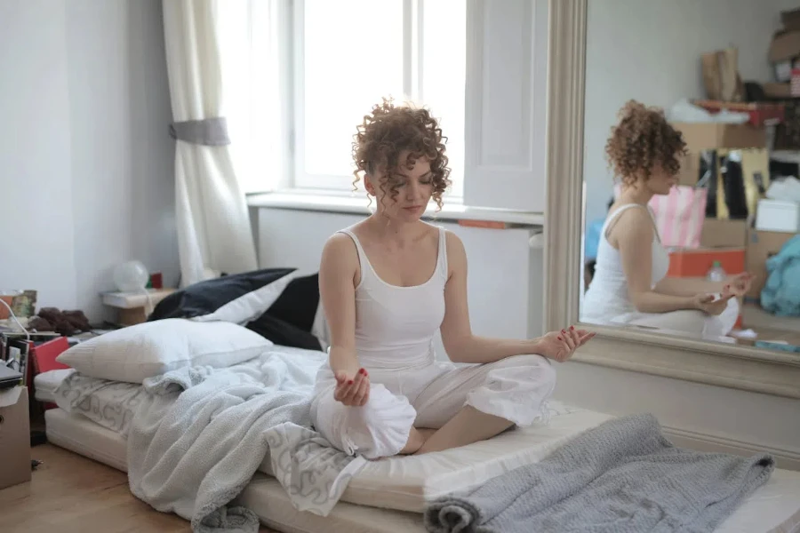 Женщина в белой пижаме медитирует на кровати.