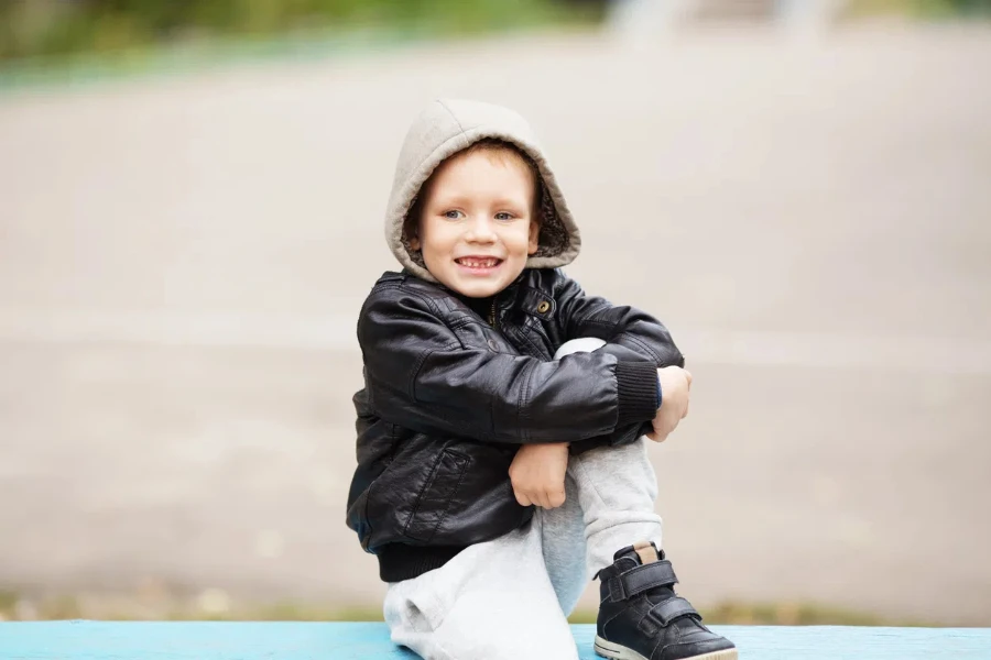 Adorable niño vestido con una chaqueta de cuero y pantalones deportivos de color gris blanquecino