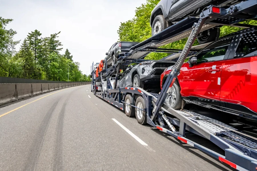 Großer industrieller Autotransporter mit Sattelzugmaschine, der Fahrzeuge auf dem modularen hydraulischen Sattelauflieger mit zwei Ebenen transportiert, der auf der breiten, geteilten Autobahnstraße mit Bäumen an der Seite fährt