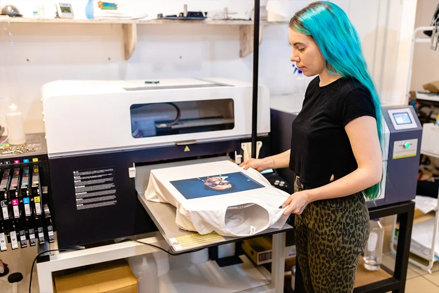 Fröhliche junge Stadtarbeiterin mit blau gefärbten Haaren bedruckt T-Shirts in der Siebdruckmaschine in ihrer Werkstatt