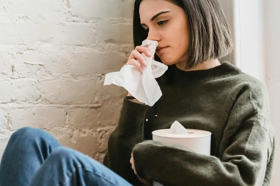 Wanita muda yang sedih dengan rambut hitam dalam sweter dengan tisu di tangan menangis dan memalingkan muka duduk di dekat dinding bata putih di rumah