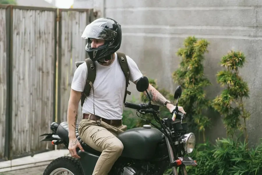 Männlicher Biker in Freizeitkleidung und Helm sitzt auf dem Motorrad und schaut im Sommer in einem Hof ​​mit geschlossenem Tor weg, von ArtHouse Studio