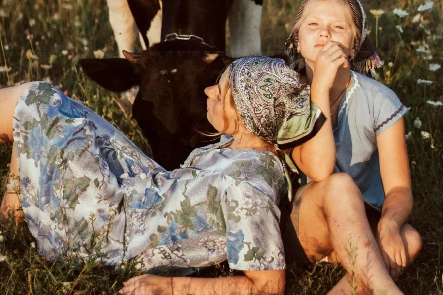 Mother and Daughter Sitting in Meadow Next to Grazing Cow