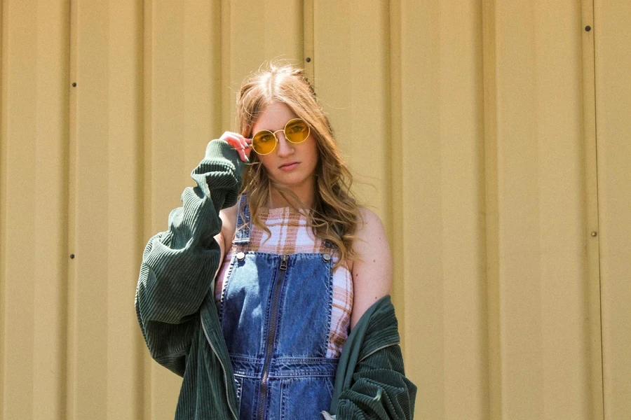 Foto de uma mulher com macacão jeans azul, jaqueta de veludo verde e óculos de sol amarelos posando