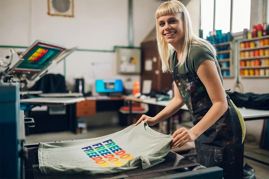 Porträt einer lächelnden Mitarbeiterin einer Druckerei, die ein im Siebdruckverfahren bedrucktes Textilprodukt in die Trocknungsmaschine der Anlage legt