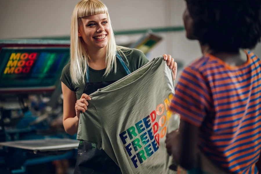 Porträt einer lächelnden Mitarbeiterin einer Druckerei, die mit einem im Siebdruckverfahren bedruckten T-Shirt in der Hand vor der Druckerei steht