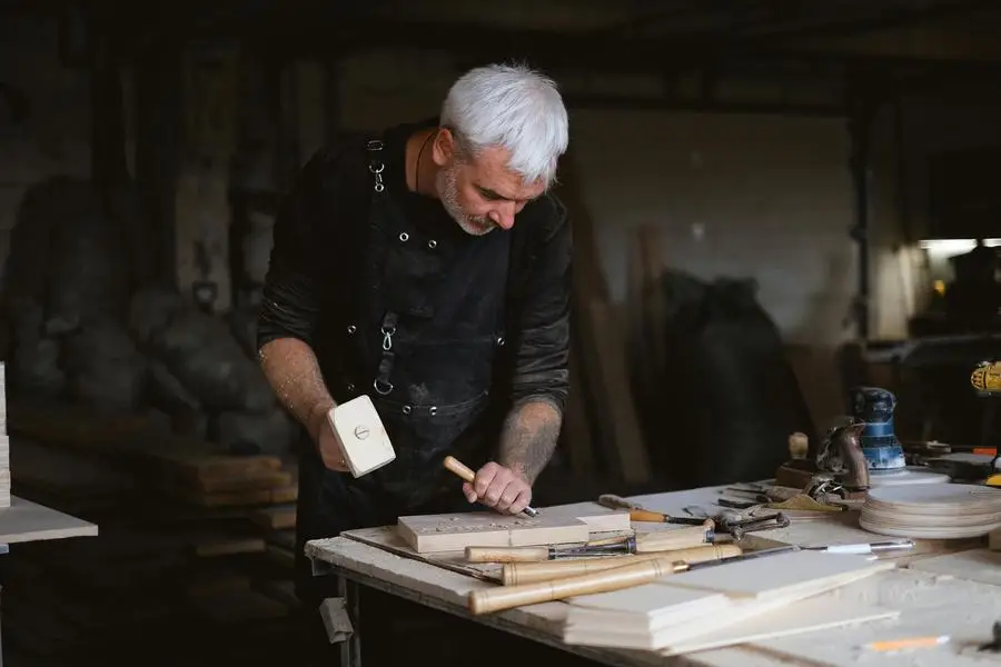 Serious male woodworker in apron using mallet and chisel to carve wooden board at table with abundance of instruments in carpentry by Anna Shvets