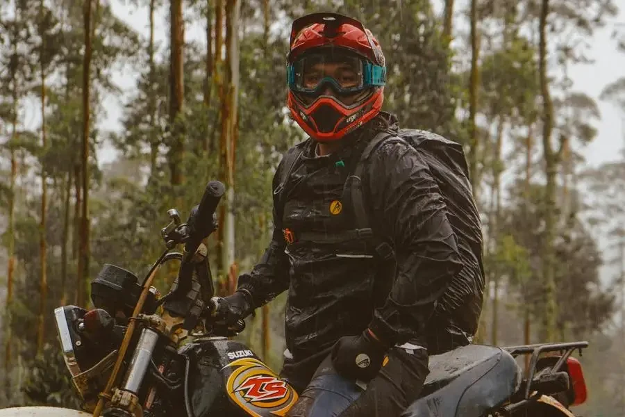 Vue latérale d'un motard masculin portant un casque de protection avec un sac à dos assis sur une moto dans les bois par Labskiii