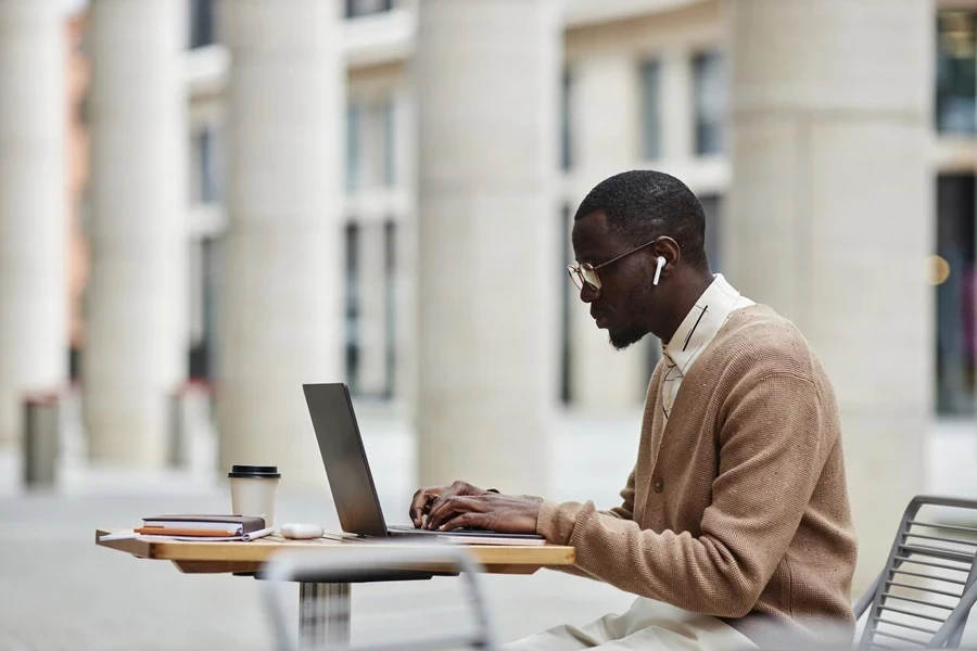 Vista lateral do jovem empresário afro-americano em fones de ouvido e roupas casuais, sentado à mesa em frente ao laptop e networking no café de rua