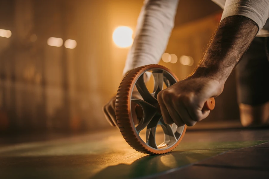 Un homme fort tenant fermement une roue abdominale pendant qu'il s'exerce seul au sol dans une salle de sport sombre