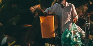 Un hombre negro irreconocible con ropa informal y gorra recoge basura de los contenedores de basura de la calle en un país tropical, por Thom Gonzalez