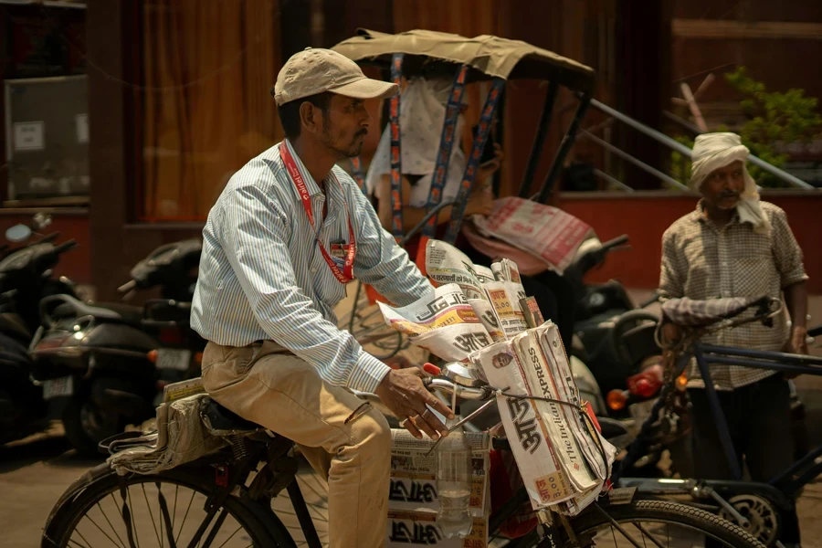 A Man Selling Newspapers Riding a Bicycle on a Street