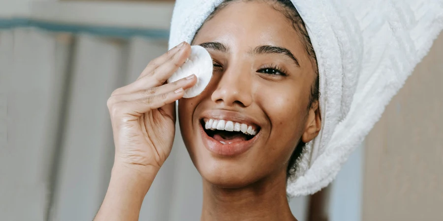 Cheerful ethnic female wrapped in towel turban moisturizing face with lotion while smiling happily