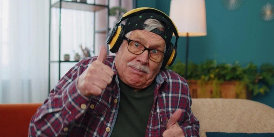 Happy senior man in wireless headphone, sitting on cozy couch in living room