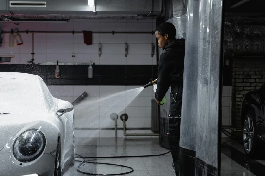 Homem de jaqueta preta lavando o carro branco usando uma mangueira de pressão de lavadora