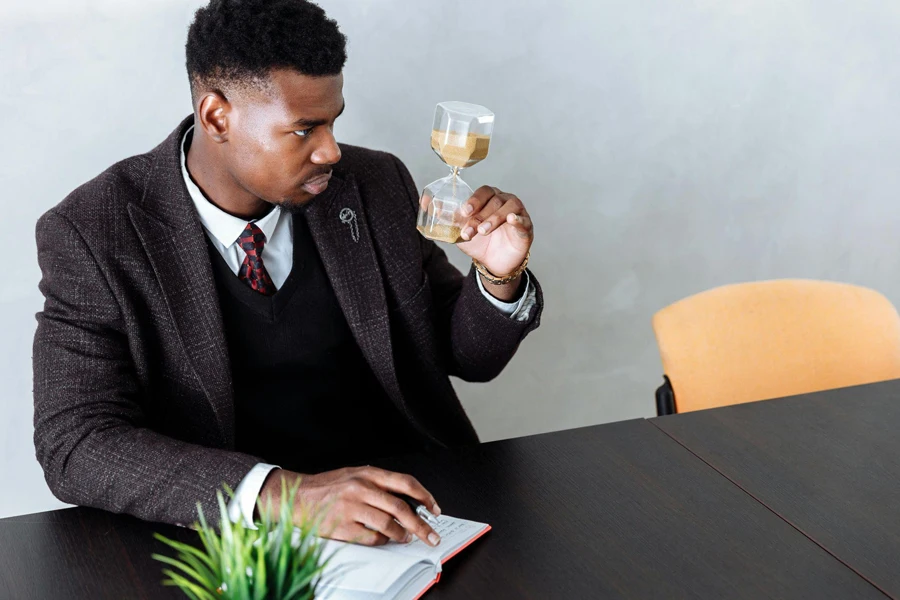 Man in Black Suit Jacket Holding and Looking at Hour Glass