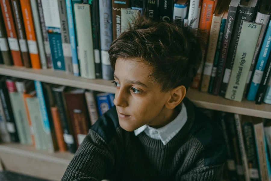 Niño con suéter sentado al lado de un estante de libros