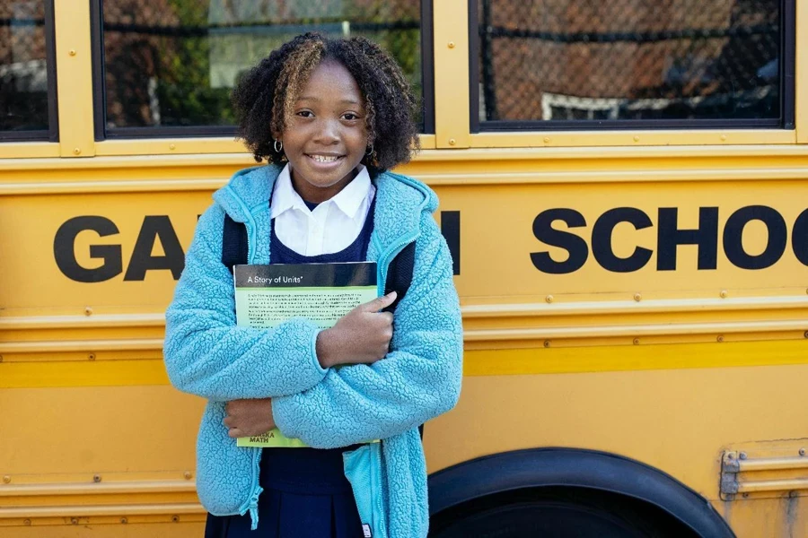 Fröhliches afroamerikanisches Mädchen in Schuluniform und warmer Jacke steht mit Lehrbuch neben einem gelben Schulbus und blickt in die Kamera