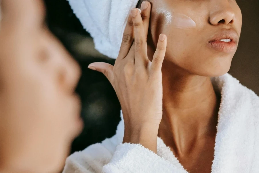 Crop Ethnic Woman Applying Cream on Face Against Mirror