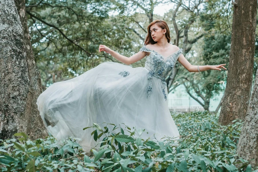 Mujer asiática joven y tranquila con un vestido romántico mirando por encima del hombro mientras baila sola en un bosque verde